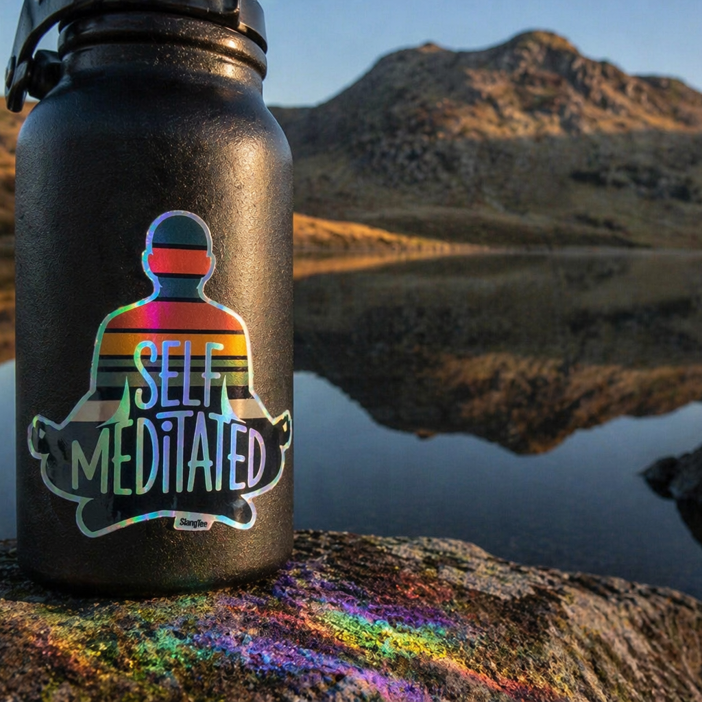 Black water bottle with 'Self Meditated' sticker on a rock by a mountain lake